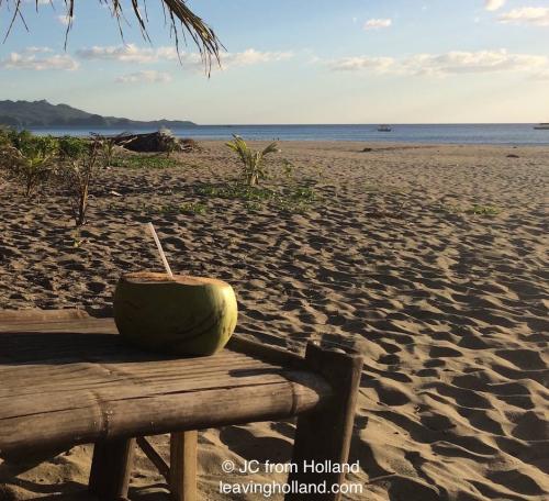 Kokosnoot op nipa tafeltje, bruin strand in licht ondergaande zon, in de verte blauwe zee