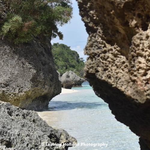 Doorkijkje tussen twee grote rotsen, wit strand, licht blauw water 