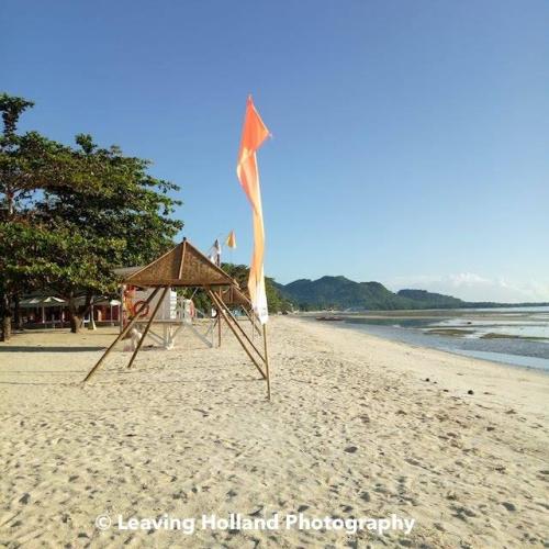 Wit strand, links bomen, rechts de zee, het is eb, op het strand oranje vlaggen en rieten parasols